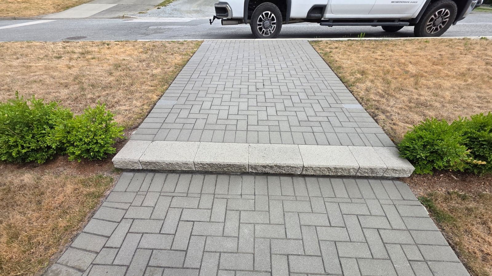 A finished interlocking paver walkway laid in a herringbone pattern on a Daedalus job, running from the street up to the house with a soldier-course border and trimmed lawn either side.
