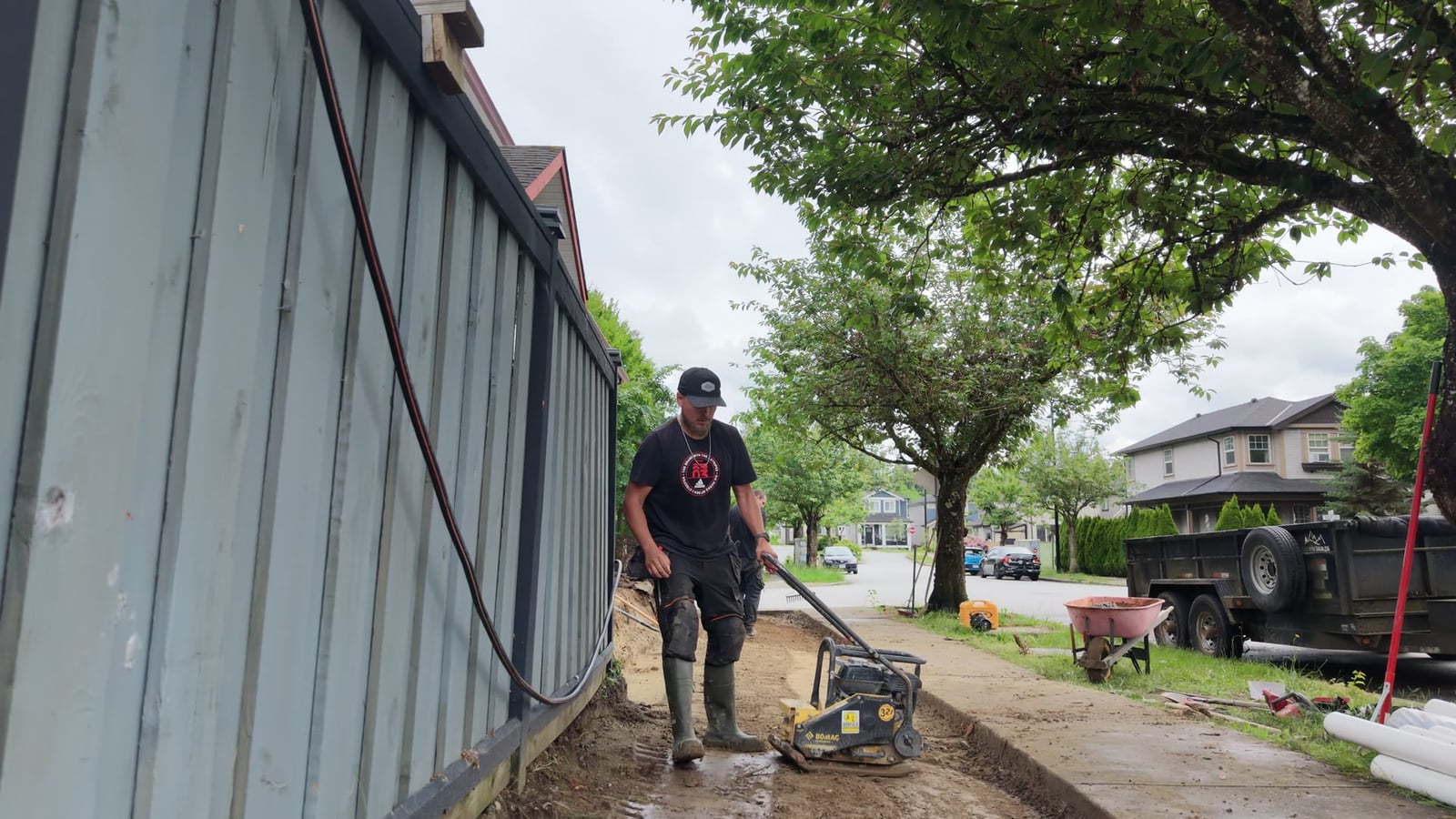 A Daedalus crew member compacting the backfill over a newly laid French drain running along the edge of a gravel drive, with the dump trailer and wheelbarrow on the drive behind.
